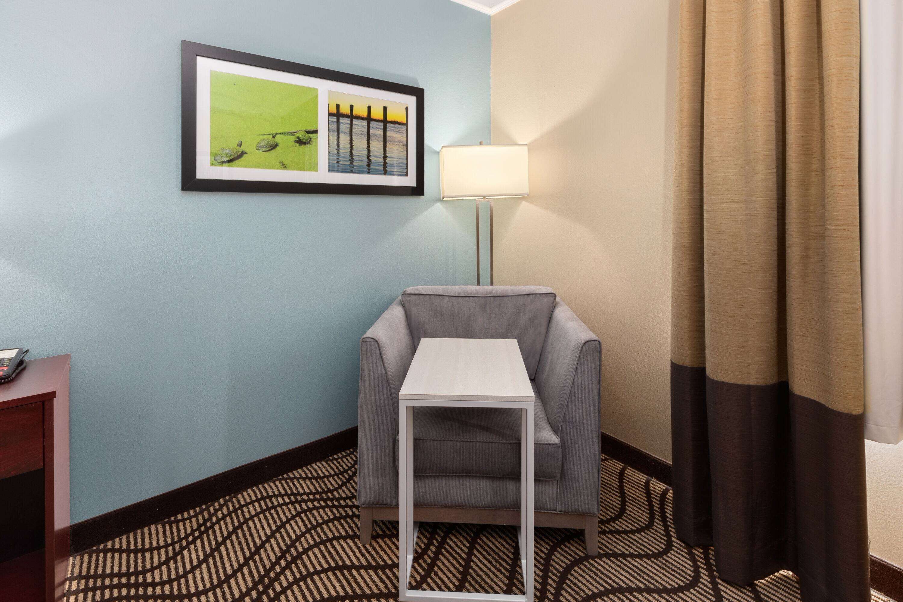 Habitación de hotel con un sillón gris, una mesita blanca pequeña, un cuadro en la pared, una lámpara de pie y una alfombra estampada.