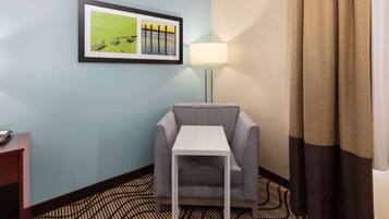 A hotel room with a grey armchair, a small white table, a framed picture on the wall, a floor lamp, and a patterned carpet.