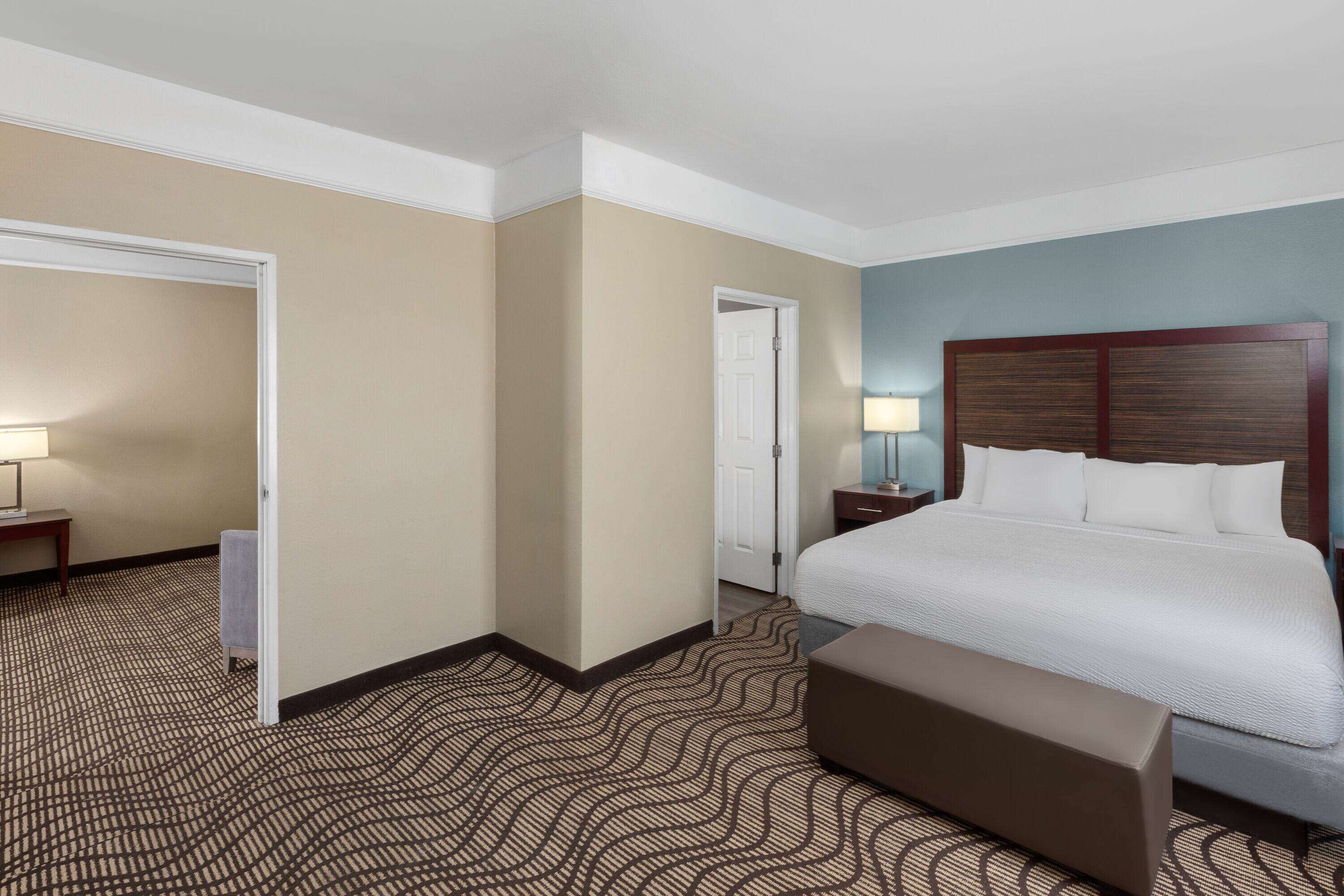Habitación de hotel con una cama grande, un banco, una mesita de noche, una lámpara y una puerta.