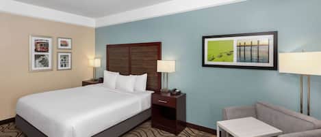 Habitación de hotel con una cama grande, una mesita de noche con lámpara, una silla y una pequeña mesa. En la pared hay cuadros enmarcados.