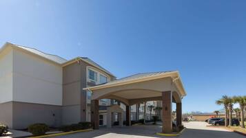 A hotel entrance with a covered walkway, parking lot, and palm trees.