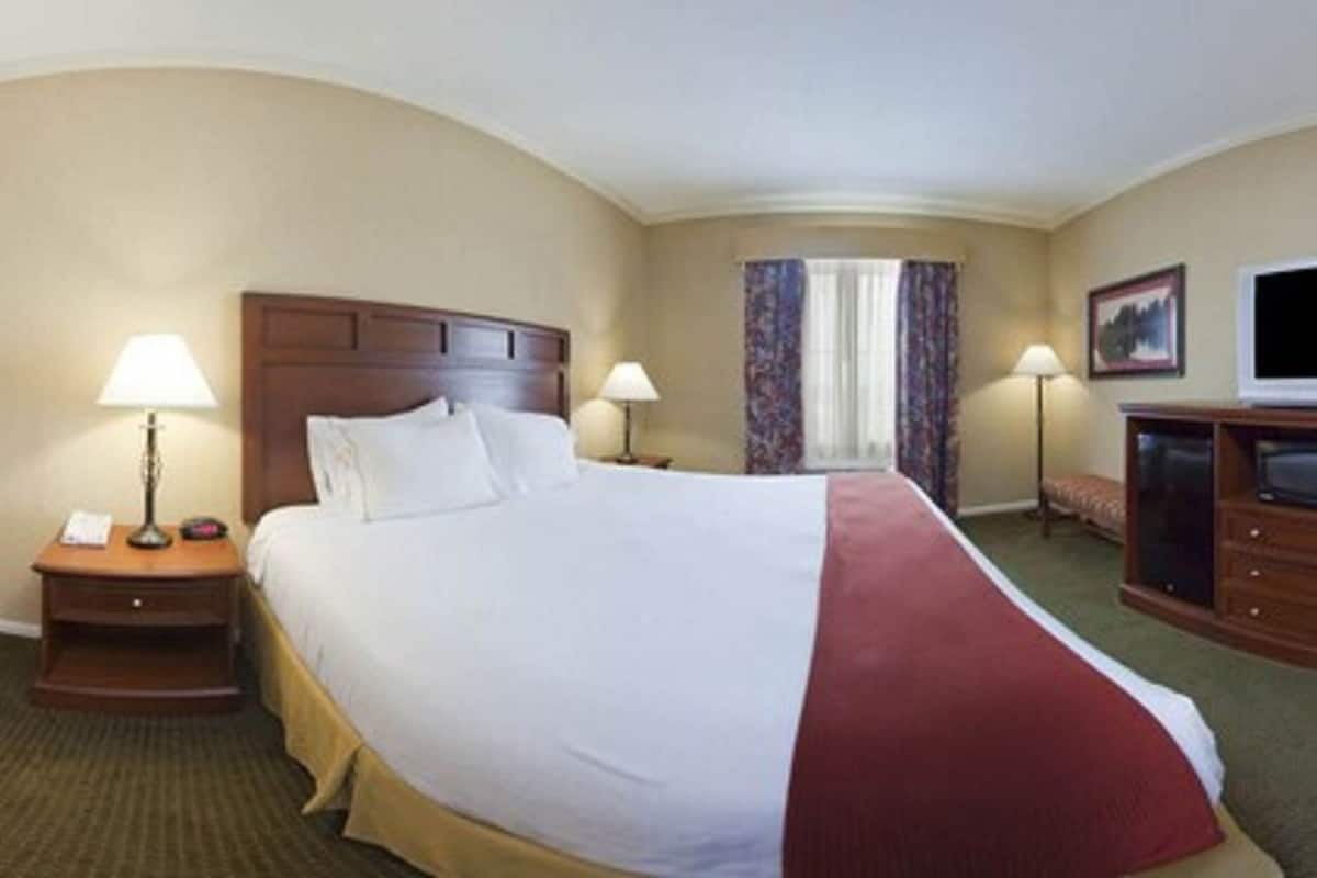 A hotel room with a large bed, two bedside tables with lamps, a television, a red armchair, and a window with curtains.