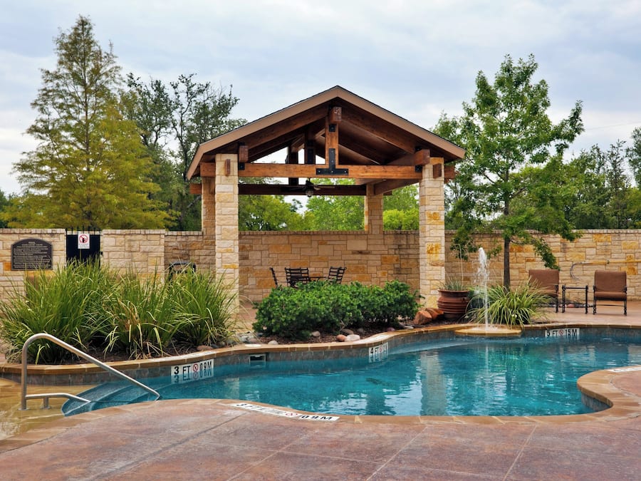 A swimming pool with a wooden gazebo, stone walls, and a fountain.