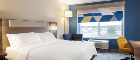 A hotel room with a bed, a desk, a chair, a window with a city view, and a wall-mounted lamp.