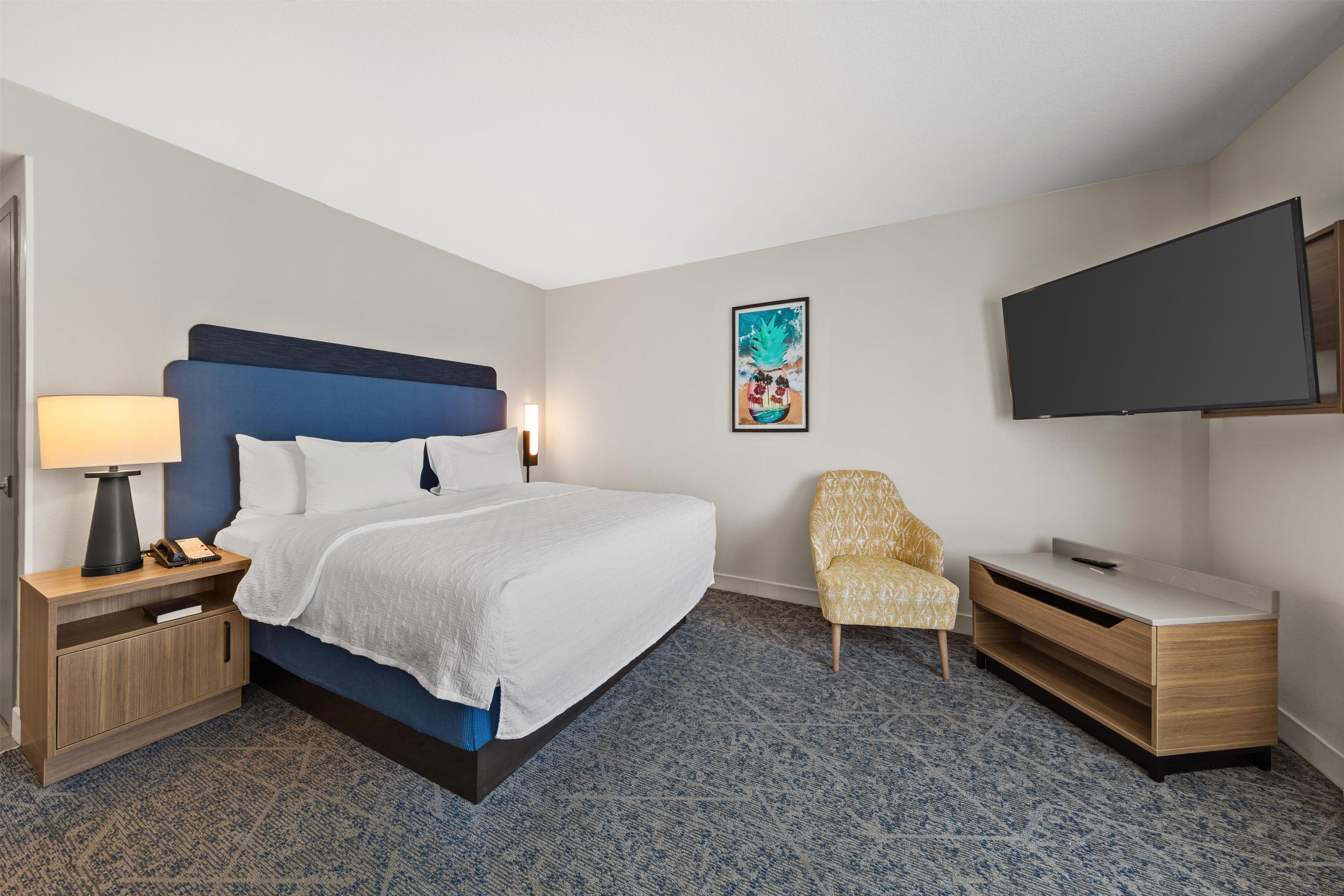 A hotel room with a bed, bedside table, lamp, chair, desk, and a TV on the wall.
