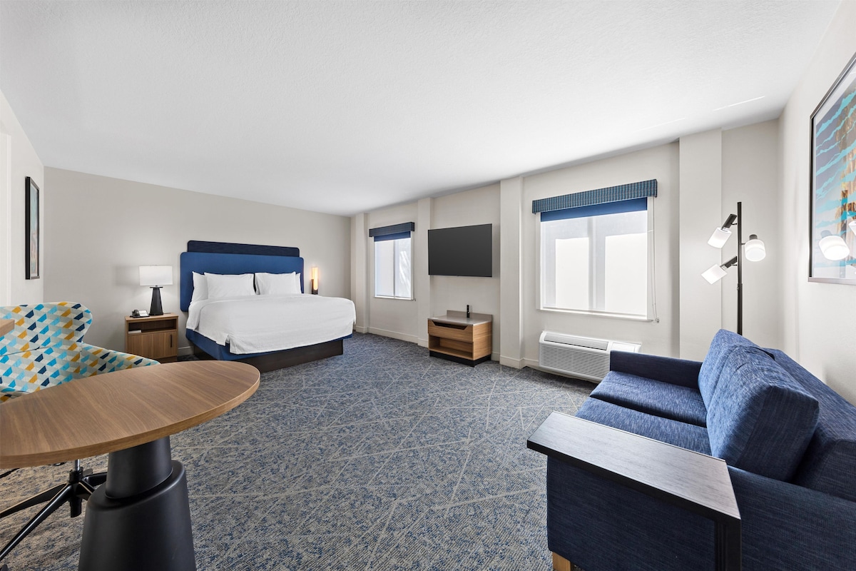 A hotel room with a bed, a desk, a chair, a sofa, a TV, and a window with curtains.