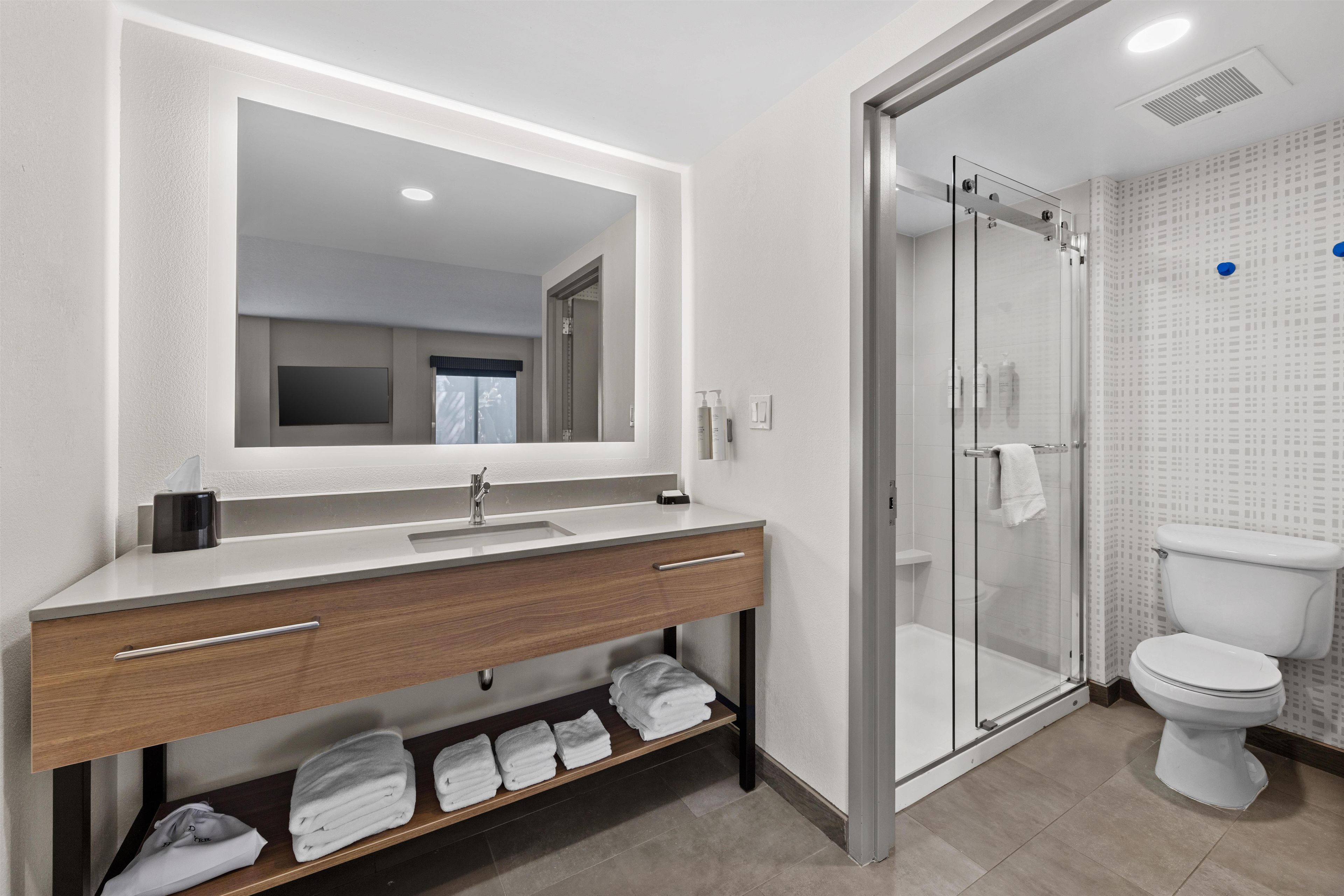 Un baño moderno con cabina de ducha de vidrio, un lavabo blanco con mueble de madera y un espejo grande.