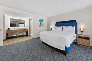A hotel room with a large bed, a wooden bench with a mirror, a nightstand with a lamp, and a framed picture on the wall.