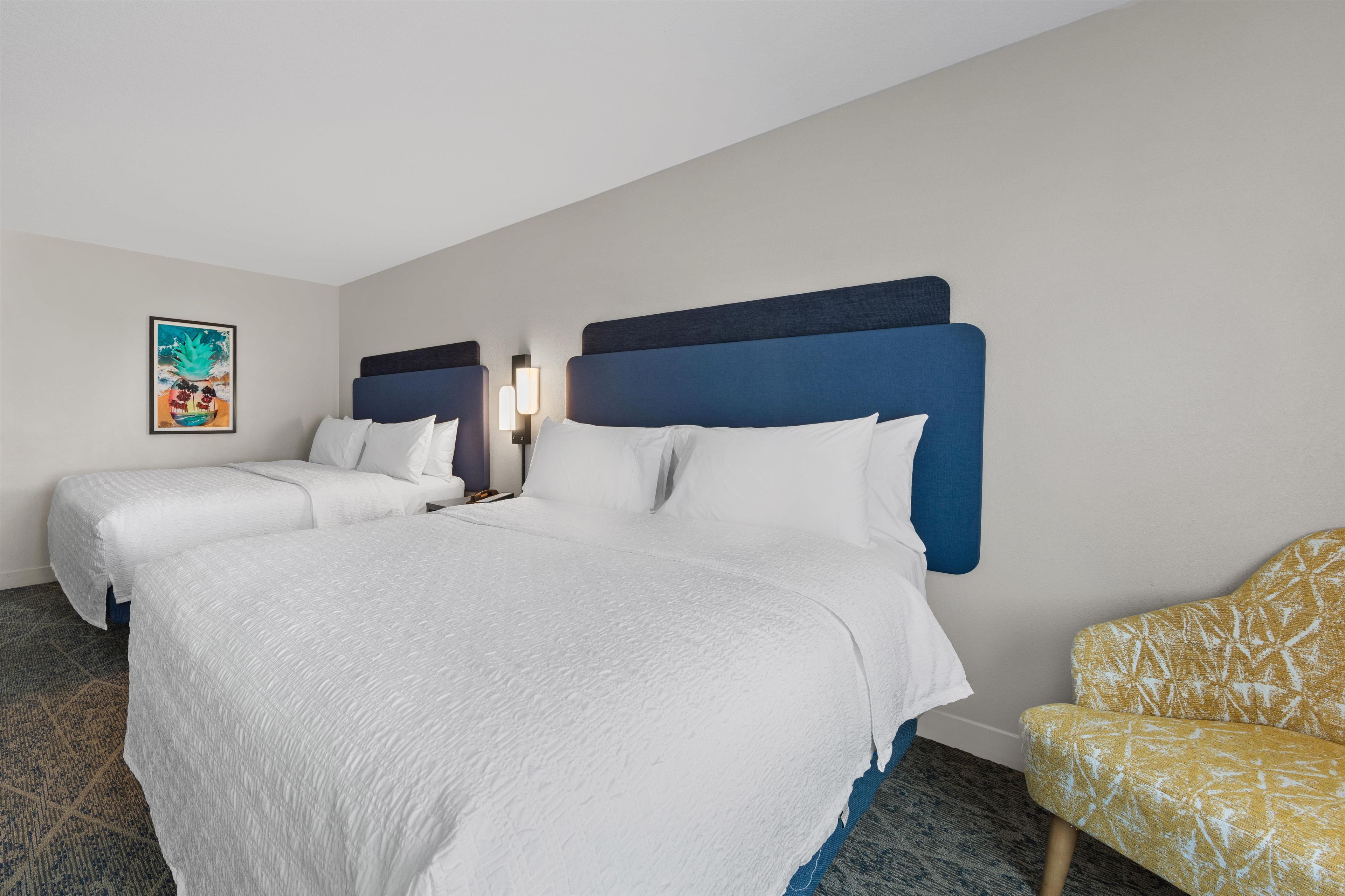 Habitación de hotel con dos camas, una mesita de noche, una lámpara, una silla y un cuadro en la pared.