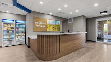 A reception desk with a 'Welcome to Hampton' sign, a counter, and two refrigerated beverage dispensers.