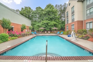 A rectangular swimming pool with a no diving sign, surrounded by a brick wall, a covered seating area with umbrellas, and a garden with various plants.
