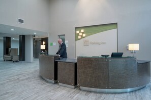 A hotel reception desk with a glass top and a sign for Holiday Inn. There are two lamps on either side of the desk, and a man is standing behind it. - Holiday Inn Atlanta/Roswell by IHG (Roswell)