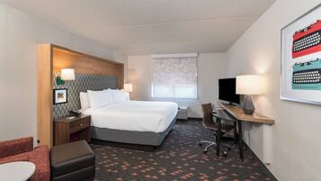 A hotel room with a bed, a desk with a computer, a chair, a TV, and a window with blinds.