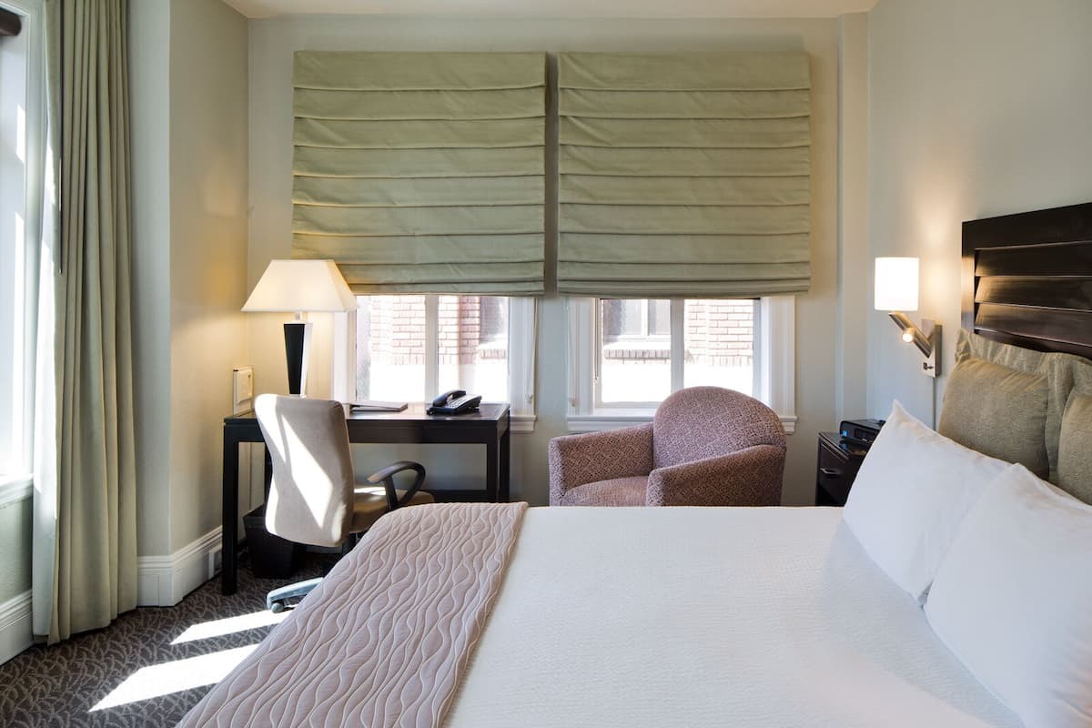 A hotel room with a large bed, a desk with a chair, a window with blinds, and a nightstand with a lamp.
