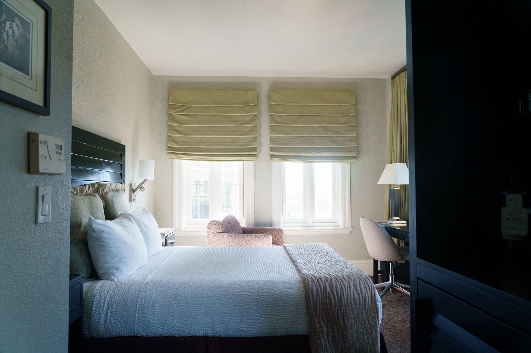 A hotel room with a bed, bedside table, lamp, and a view of the city through large windows.