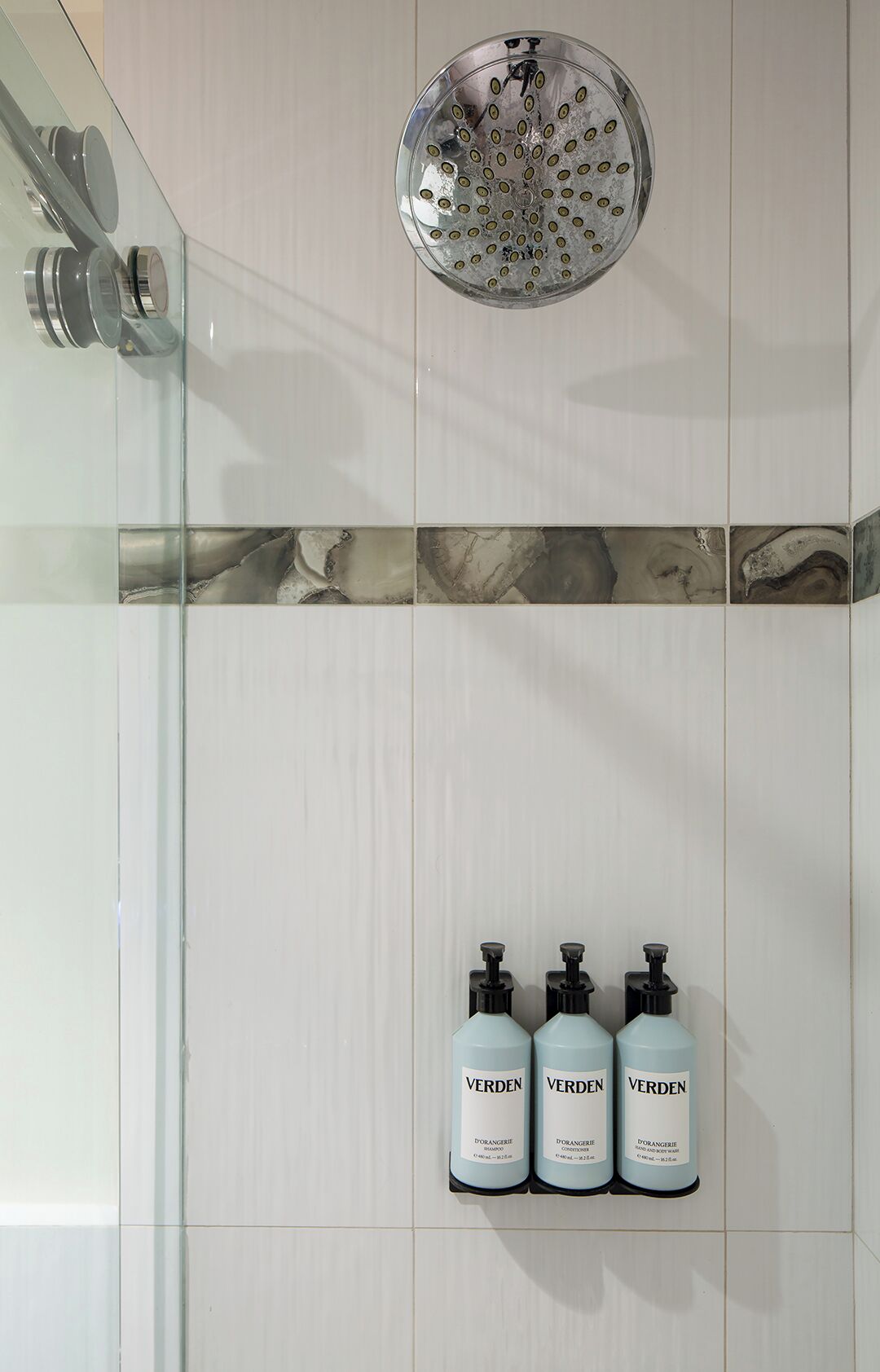 Een badkamer met een glazen douchecabine, een backsplash die op marmer lijkt, en drie flessen Verden shampoo en conditioner.