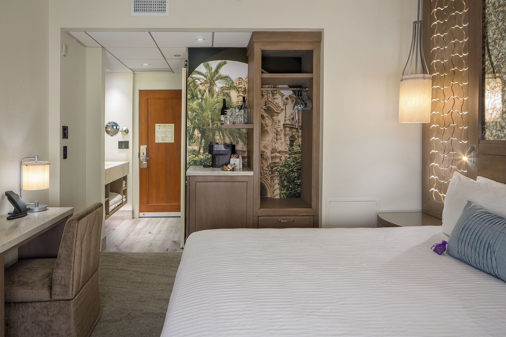 Habitación de hotel con cama, escritorio con lámpara, armario y vista a un edificio a través de una ventana.