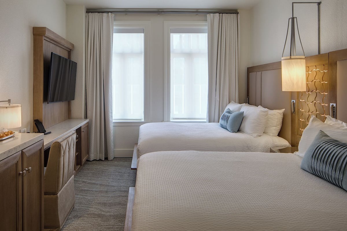 A hotel room with two beds, a desk with a lamp, a TV, and a window with curtains.