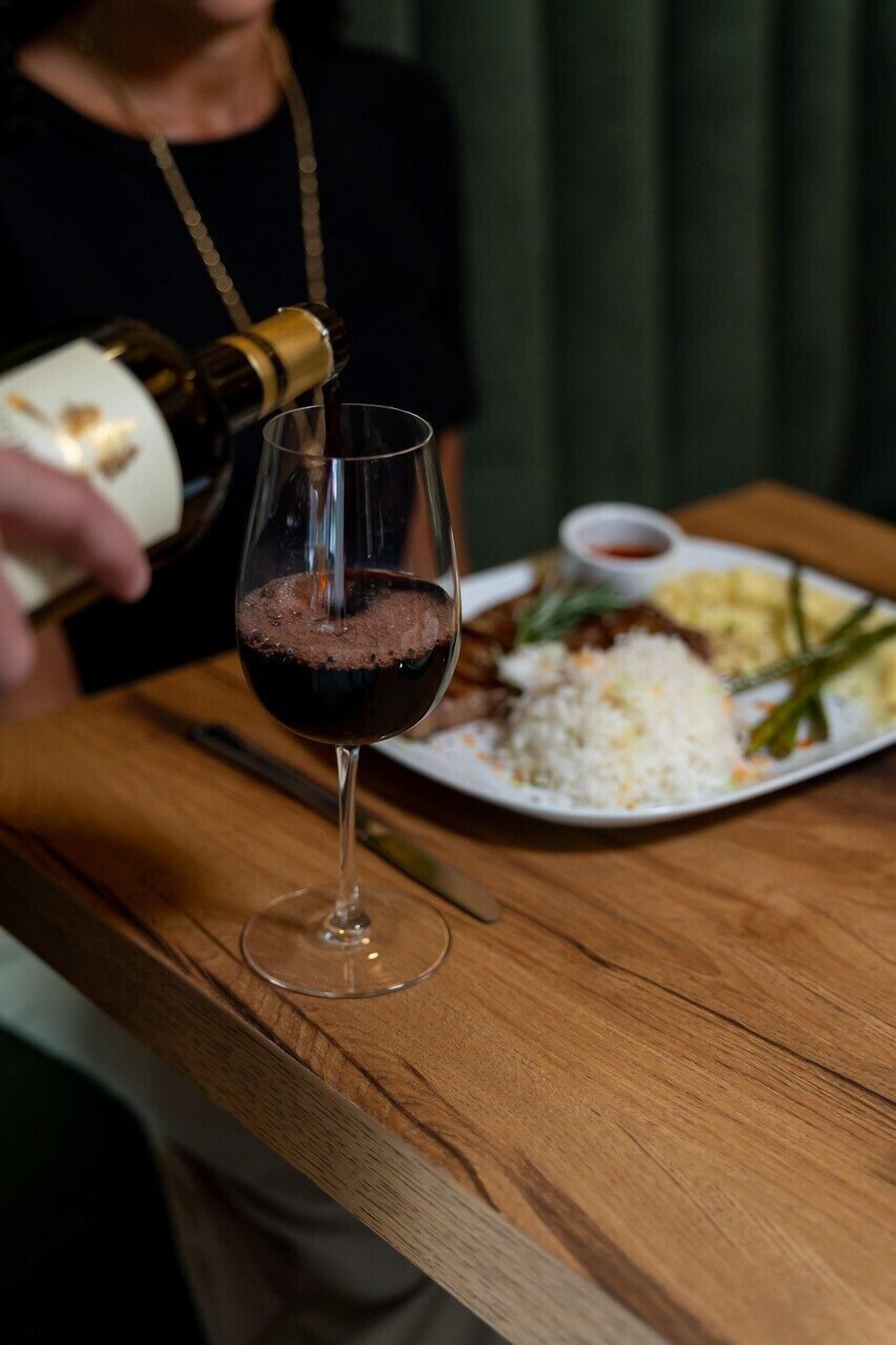 A person pouring a drink into a glass on a wooden table with a plate of food beside it.