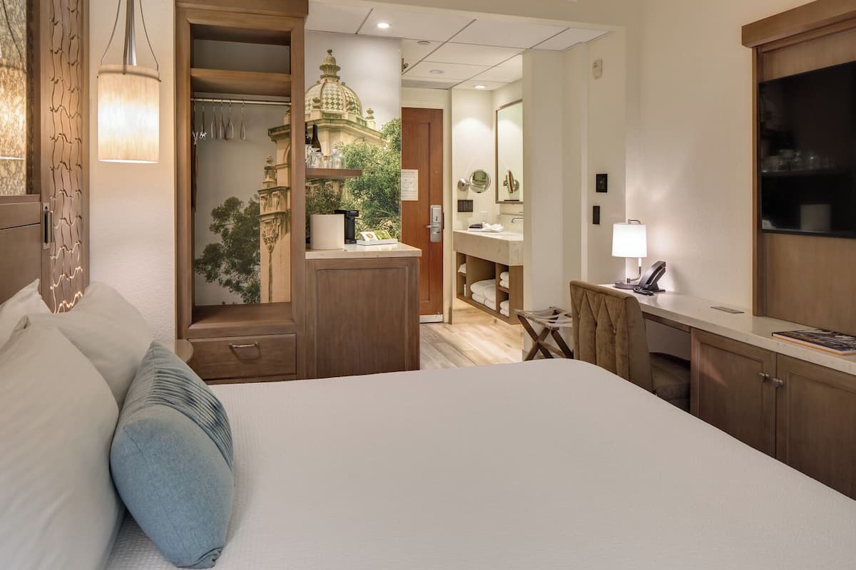 A hotel room with a large bed, a desk with a lamp, a vanity with a mirror, and a view of a temple through an open door.