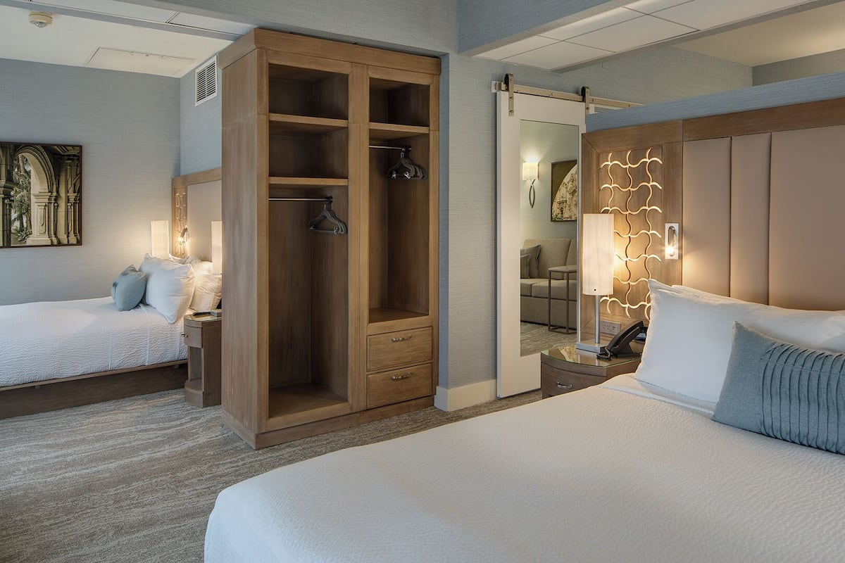 A hotel room with a large walk-in closet, a bed with white bedding, a bedside table with a lamp, and a wall-mounted mirror.