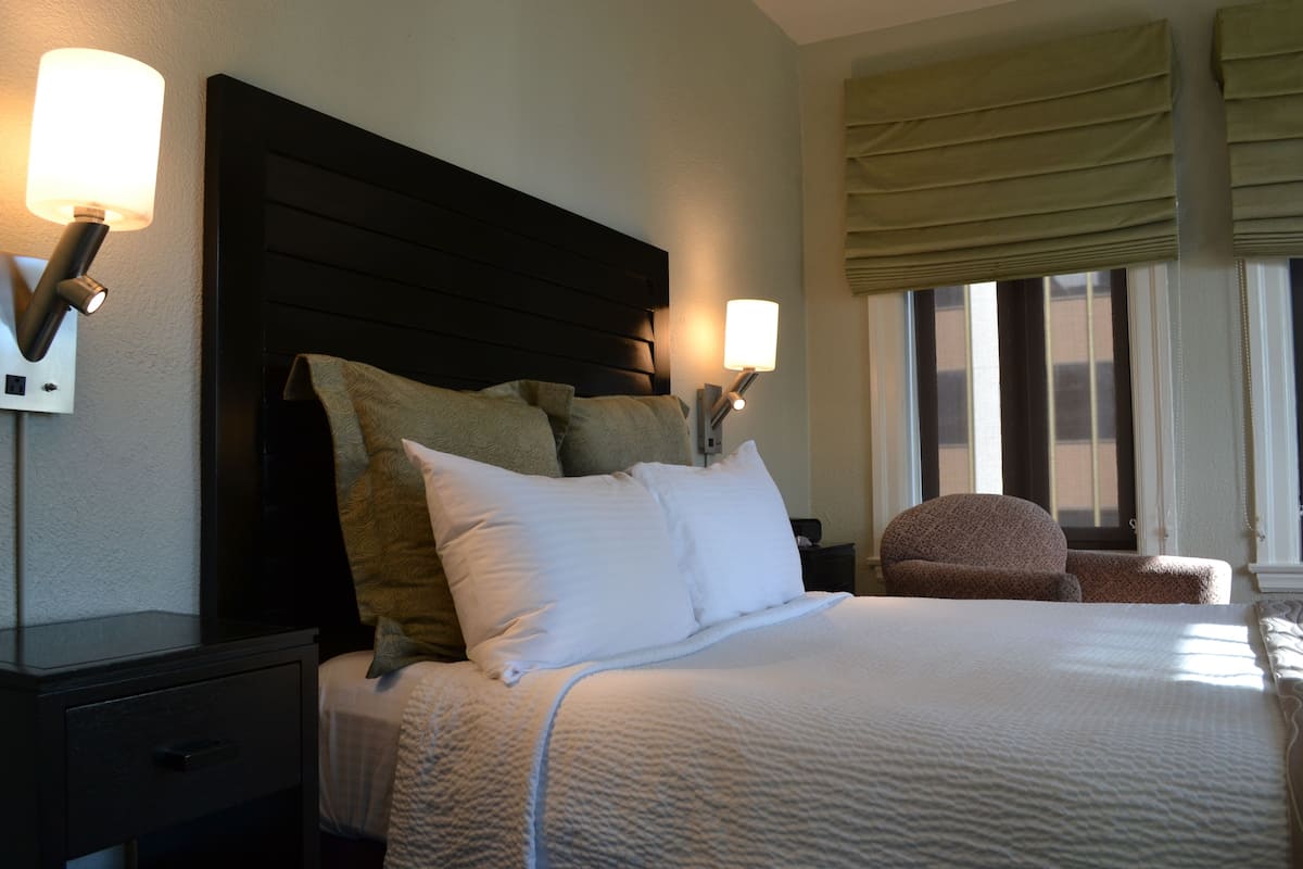 A neatly made bed with a dark headboard, two bedside lamps, and a window with curtains.