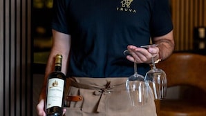 Una persona sosteniendo una botella y copas, vestida con una camisa oscura con un logo y un delantal.