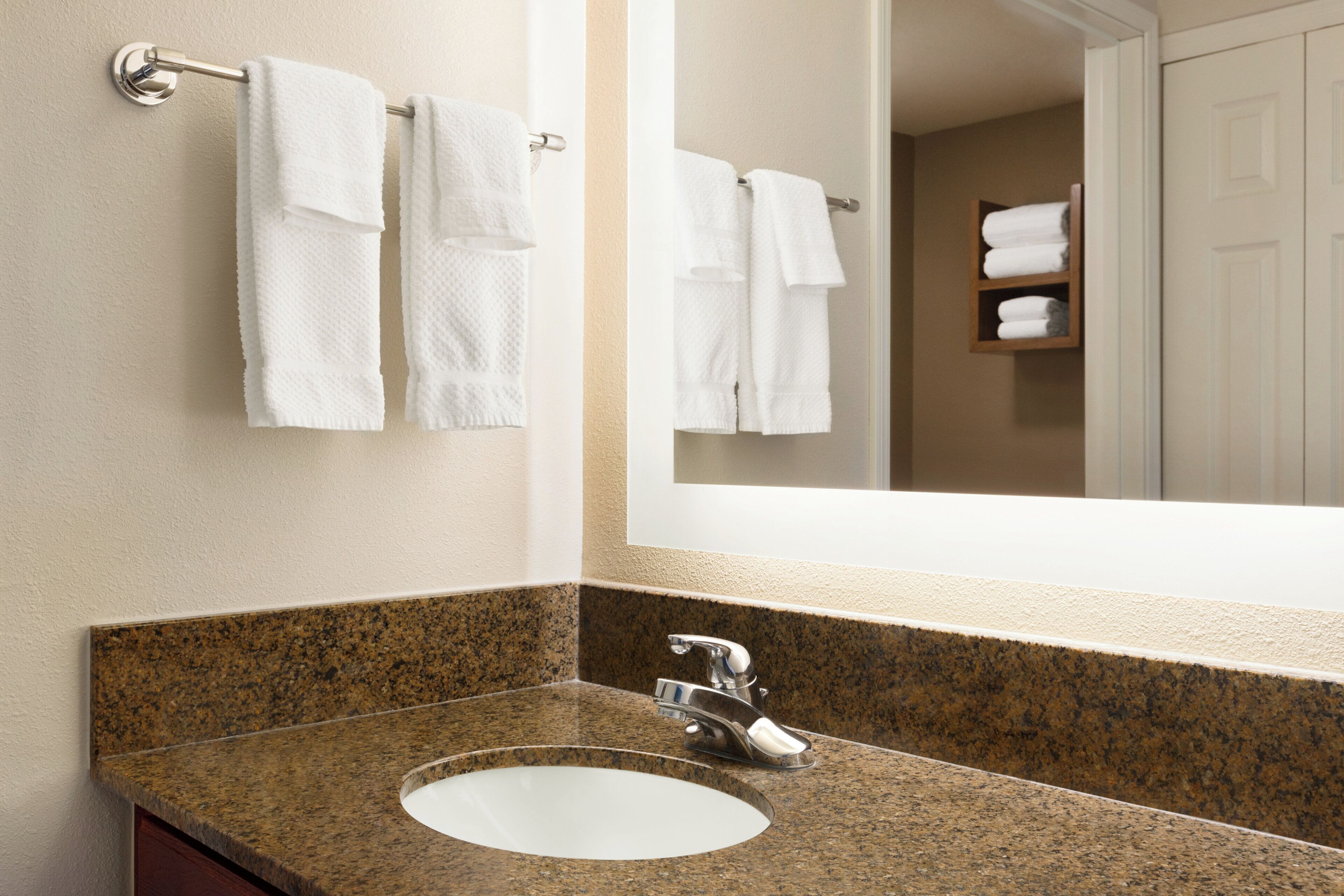 Un baño con encimera de granito, un lavamanos blanco y un espejo que refleja un perchero y una puerta.