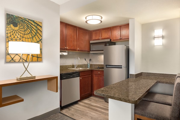 A compact kitchen with wooden cabinets, a stainless steel refrigerator, a built-in microwave, and a granite countertop.