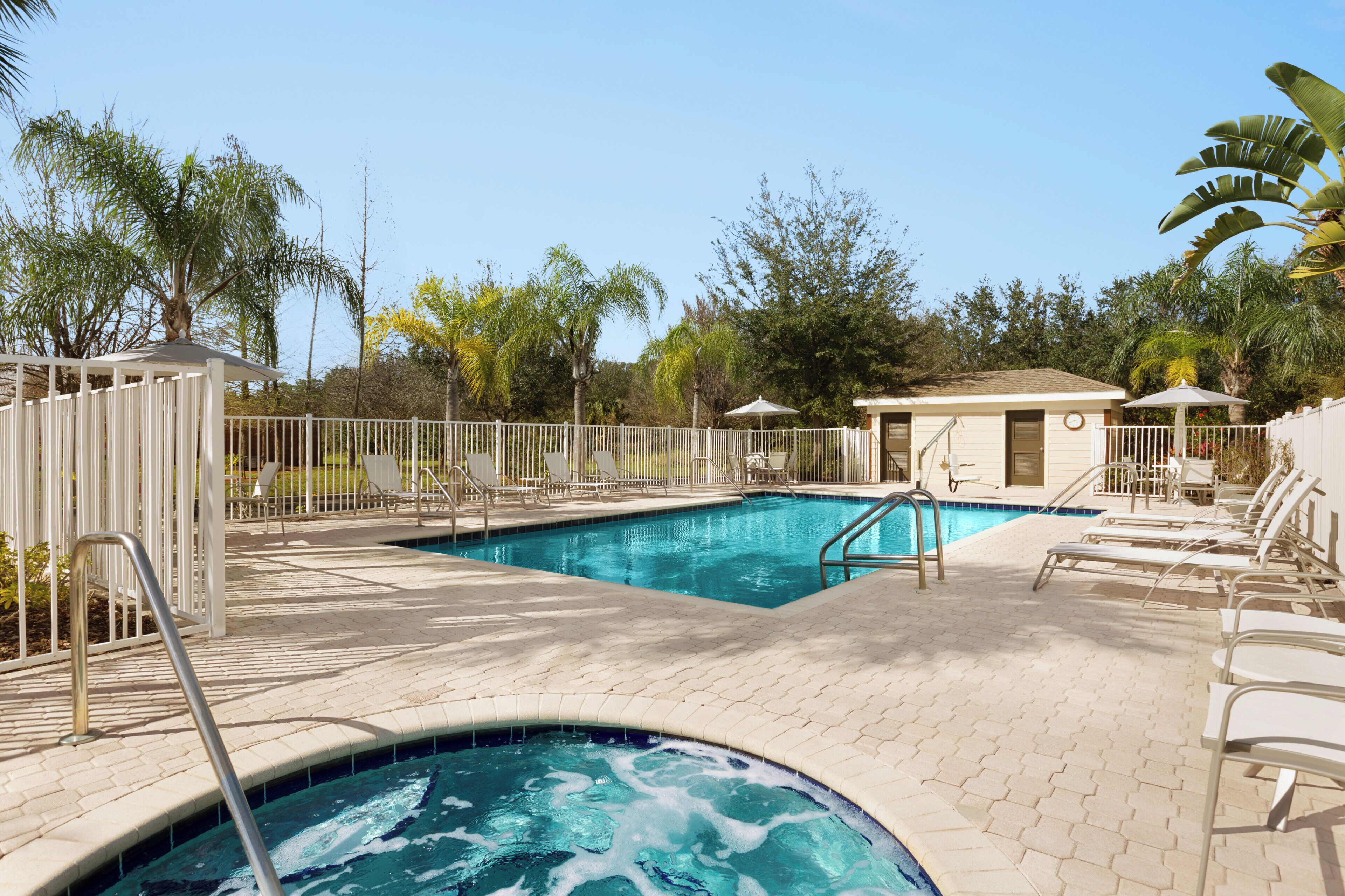 A swimming pool area with a hot tub, lounge chairs, and a small building, surrounded by palm trees.