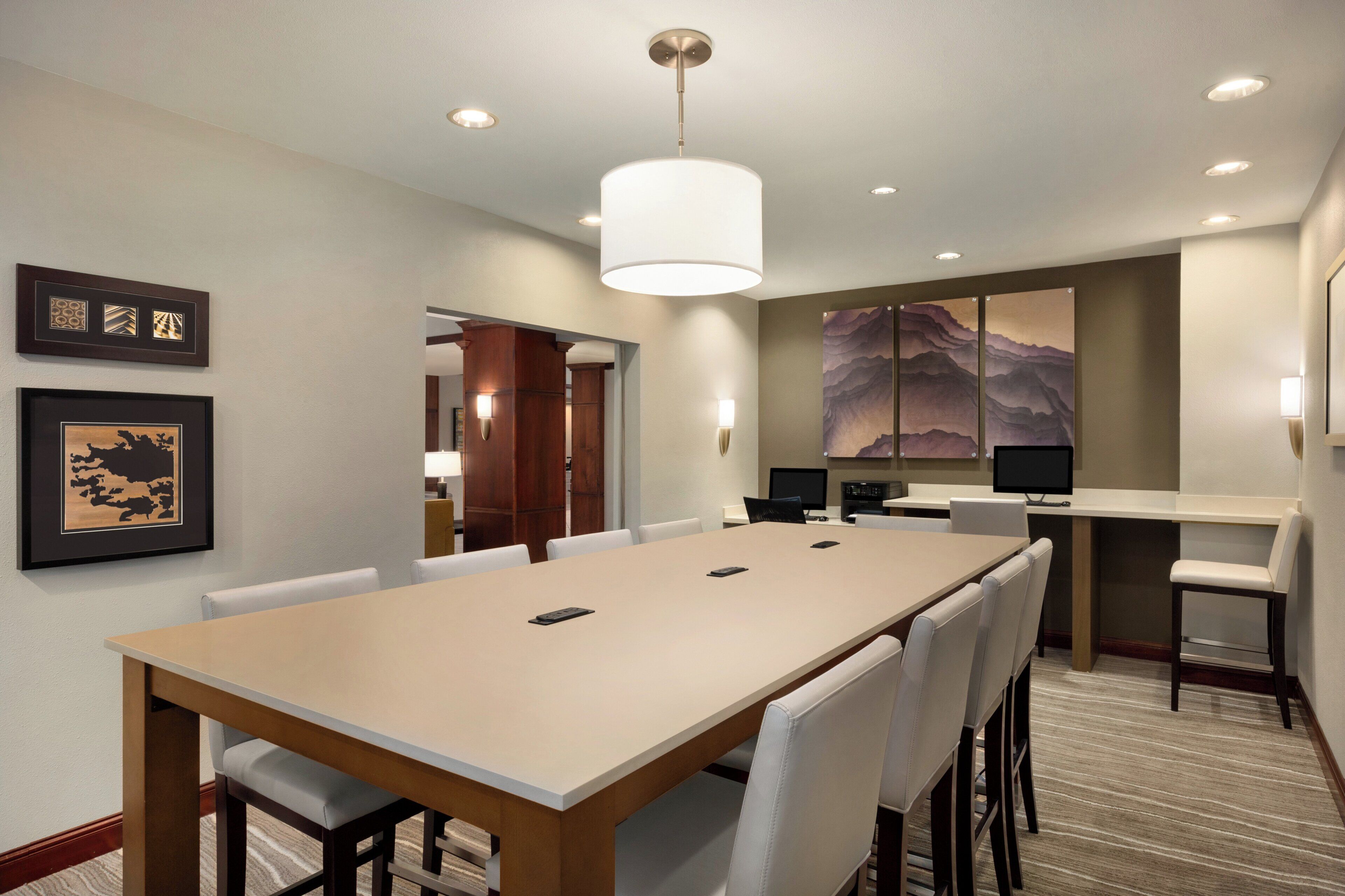 A hotel conference room with a long table, chairs, a large painting, and a computer on a desk.