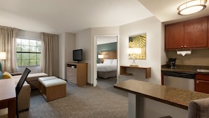 Habitación de hotel con cama, escritorio, silla, televisor, kitchenette y ventana con cortinas.