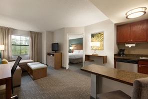A hotel room with a bed, a desk, a chair, a TV, a small kitchen area, and a window with curtains.