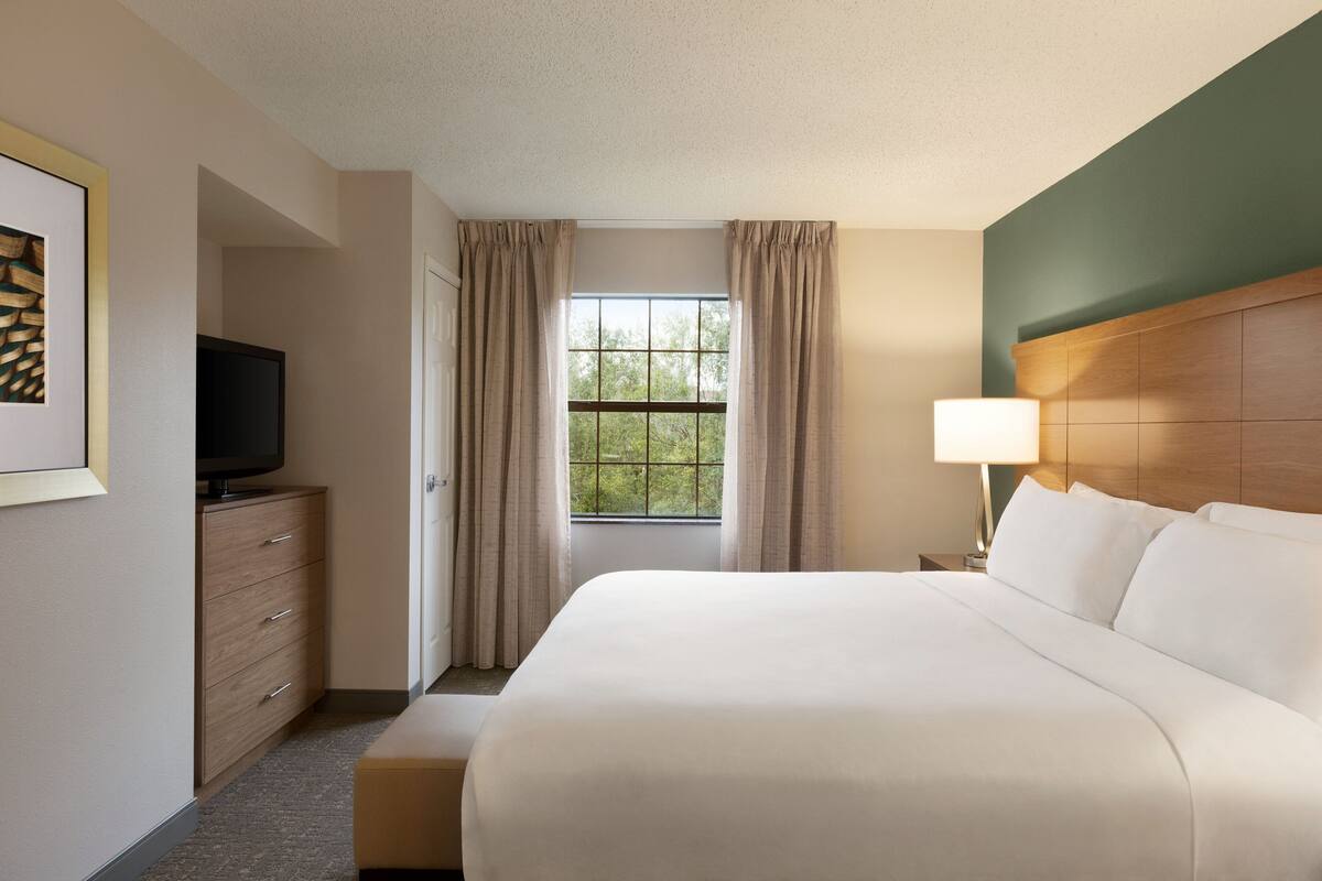 Une chambre d’hôtel avec un grand lit, un téléviseur posé sur une commode en bois, une fenêtre avec des rideaux et une table de chevet avec une lampe.