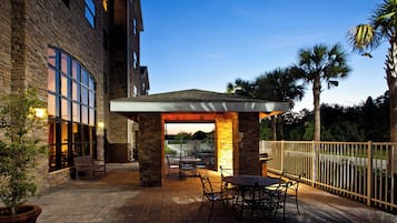 Un patio exterior con suelo de ladrillo, pilares de piedra y una zona cubierta con mesas y sillas.