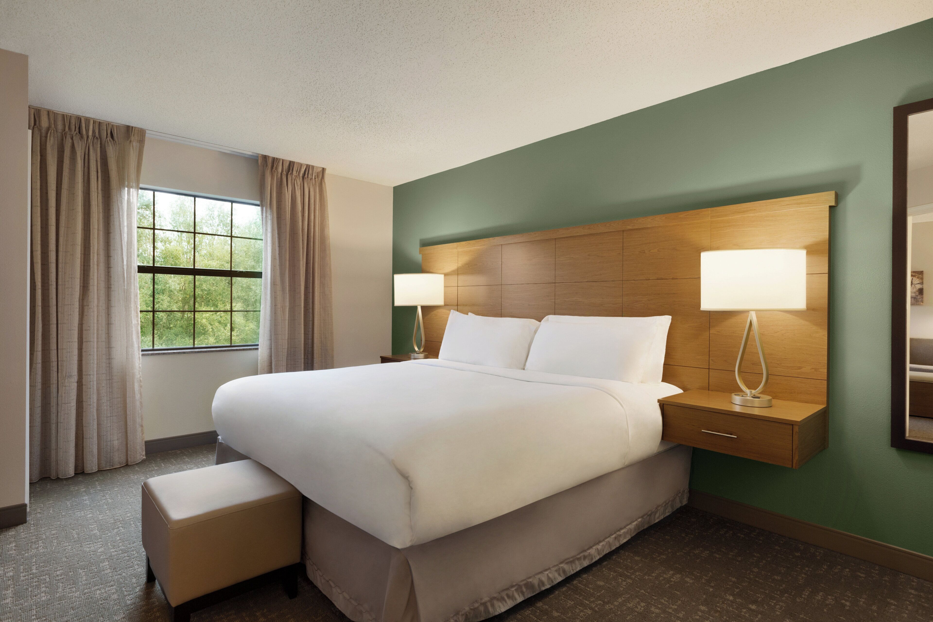 Quarto de hotel com uma cama grande, duas mesas de cabeceira, um criado-mudo, janela com cortinas e parede verde.