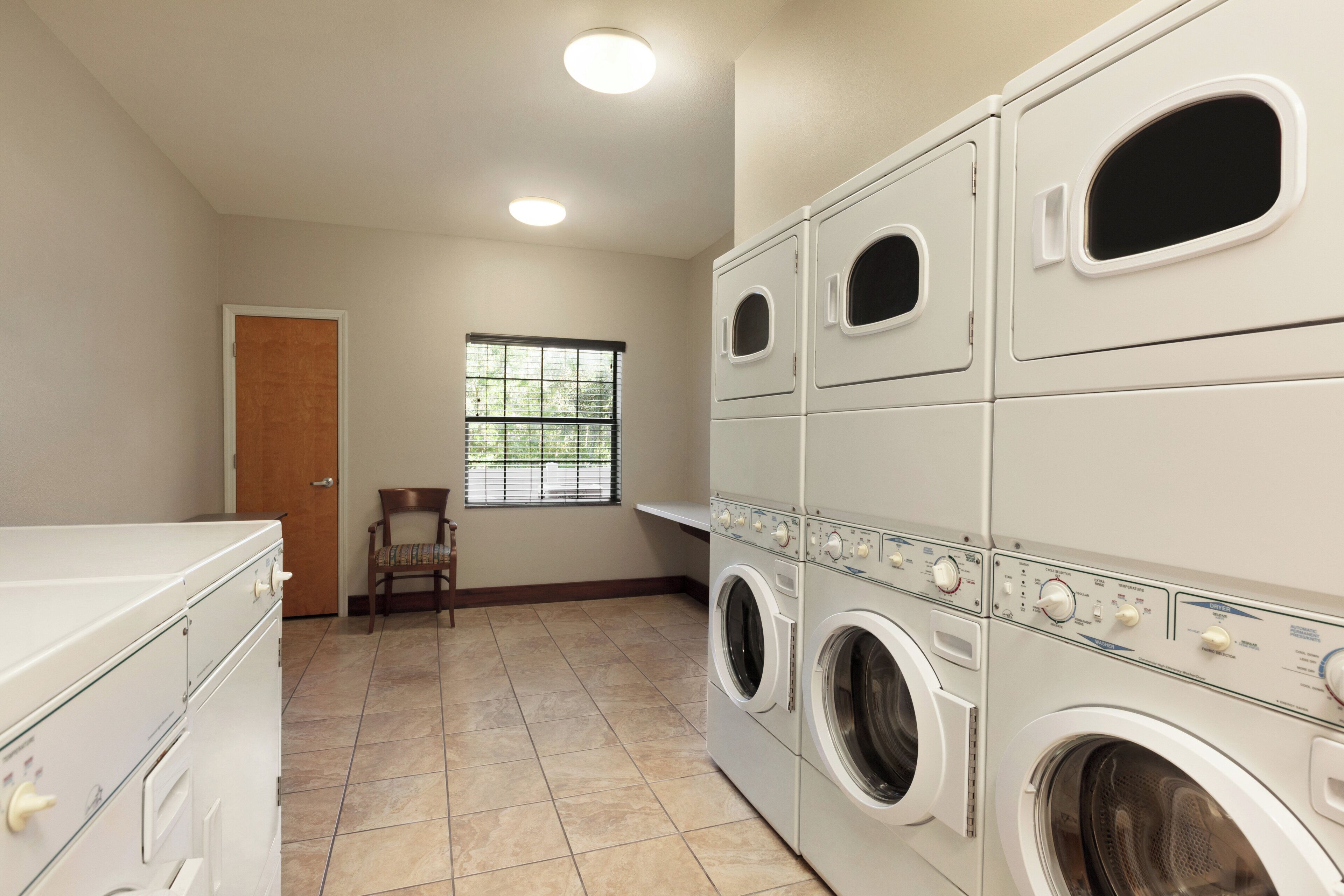 A laundry room with stacked washers and dryers, a wooden chair, a door, and a window with blinds.