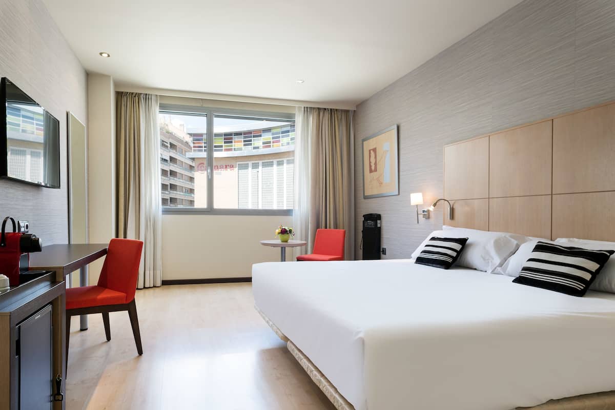 Habitación de hotel con una cama, un escritorio, una silla, una mesita con un florero y una ventana con cortinas.