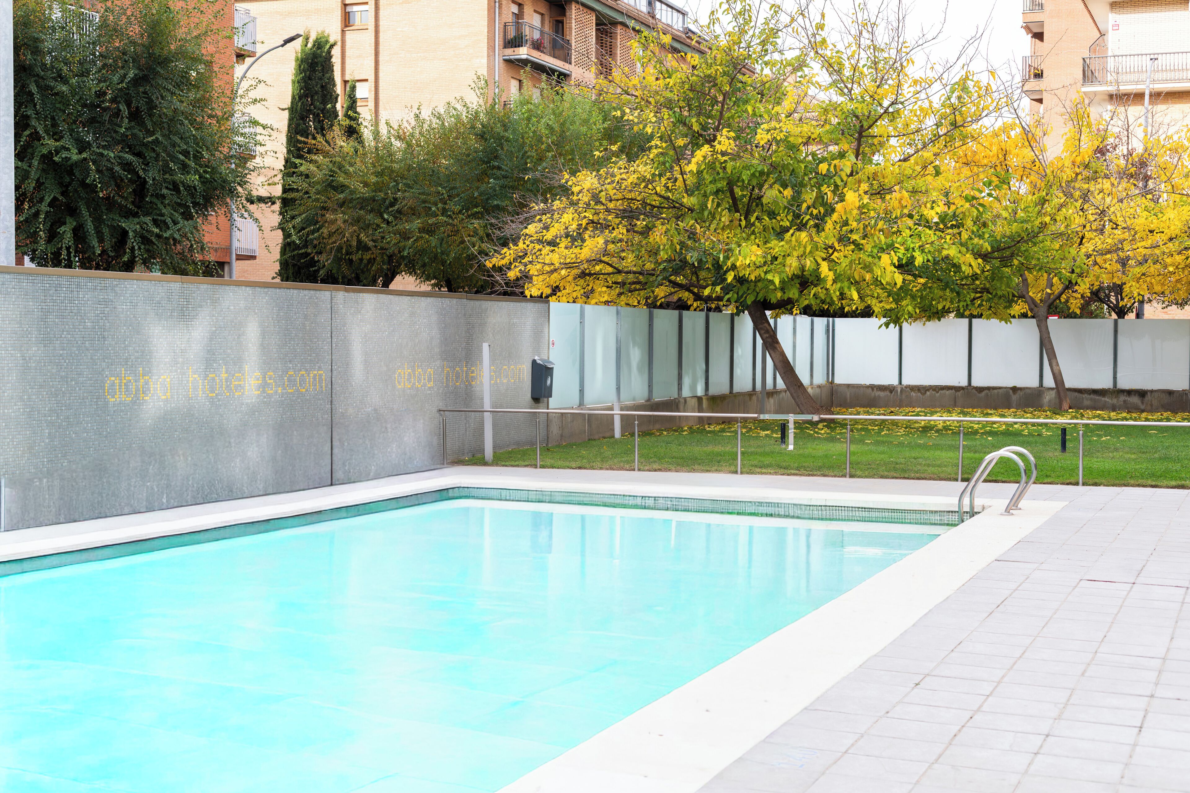Ein Schwimmbecken mit gefliestem Deck, umgeben von einer niedrigen Mauer und einem Baum mit gelben Blättern.