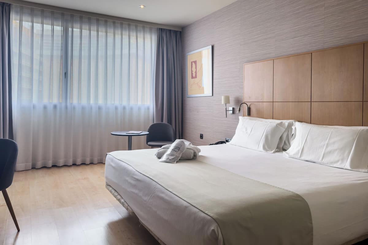 Habitación de hotel con una cama grande, una mesita pequeña, una silla, una lámpara de pared y un ventanal con cortinas.
