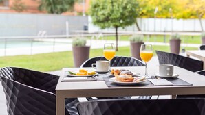 A dining table with breakfast items and two glasses of orange juice, set against a backdrop of a pool and greenery.