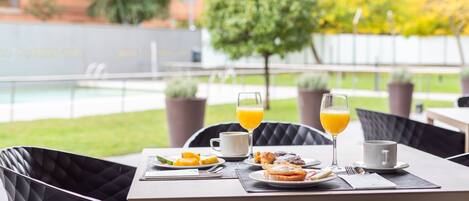 A dining table with breakfast items and two glasses of orange juice, set against a backdrop of a pool and greenery.
