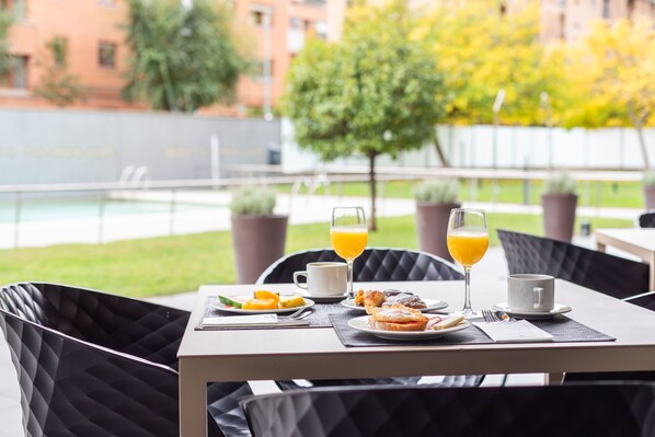 A dining table with breakfast items and two glasses of orange juice, set against a backdrop of a pool and greenery.