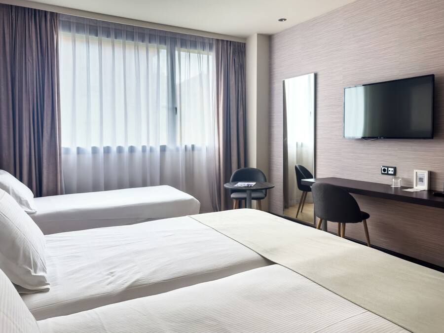 Quarto de hotel com duas camas, uma mesa, uma cadeira, uma televisão e uma janela com cortinas.