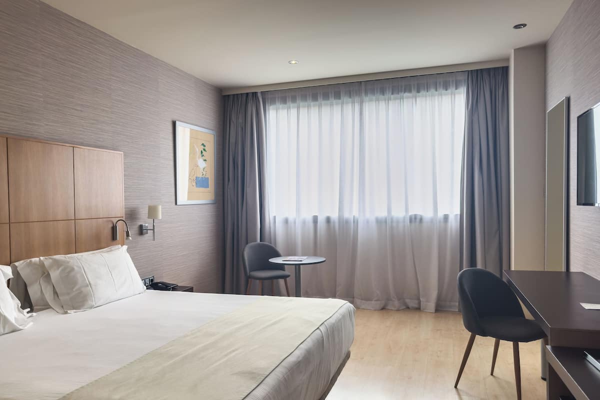 Habitación de hotel con cama, escritorio, silla, mesita, lámpara, un cuadro y un ventanal con cortinas.