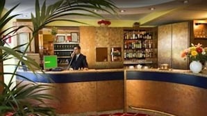 A reception area with a counter, a man standing behind it, and a bar with shelves of bottles. - Cinque Giornate (Milan)