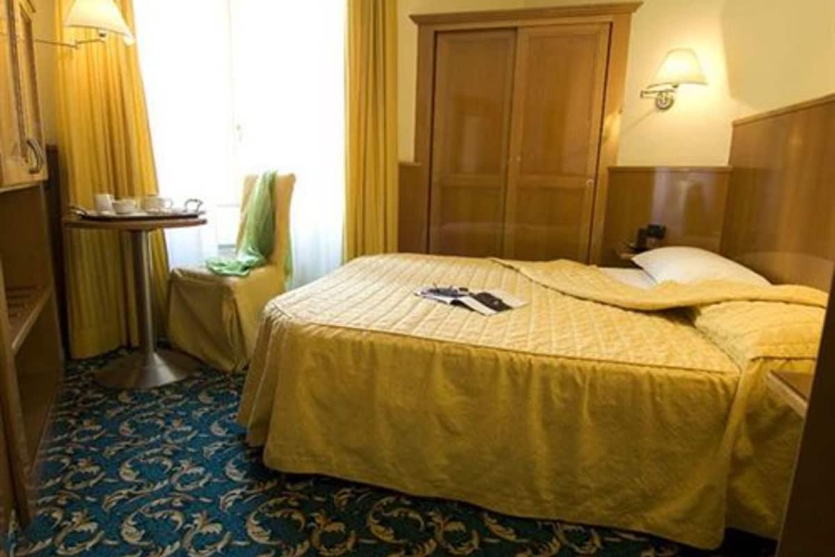 A hotel room with a bed, a wooden wardrobe, a small table with a tray of items, and a lamp.