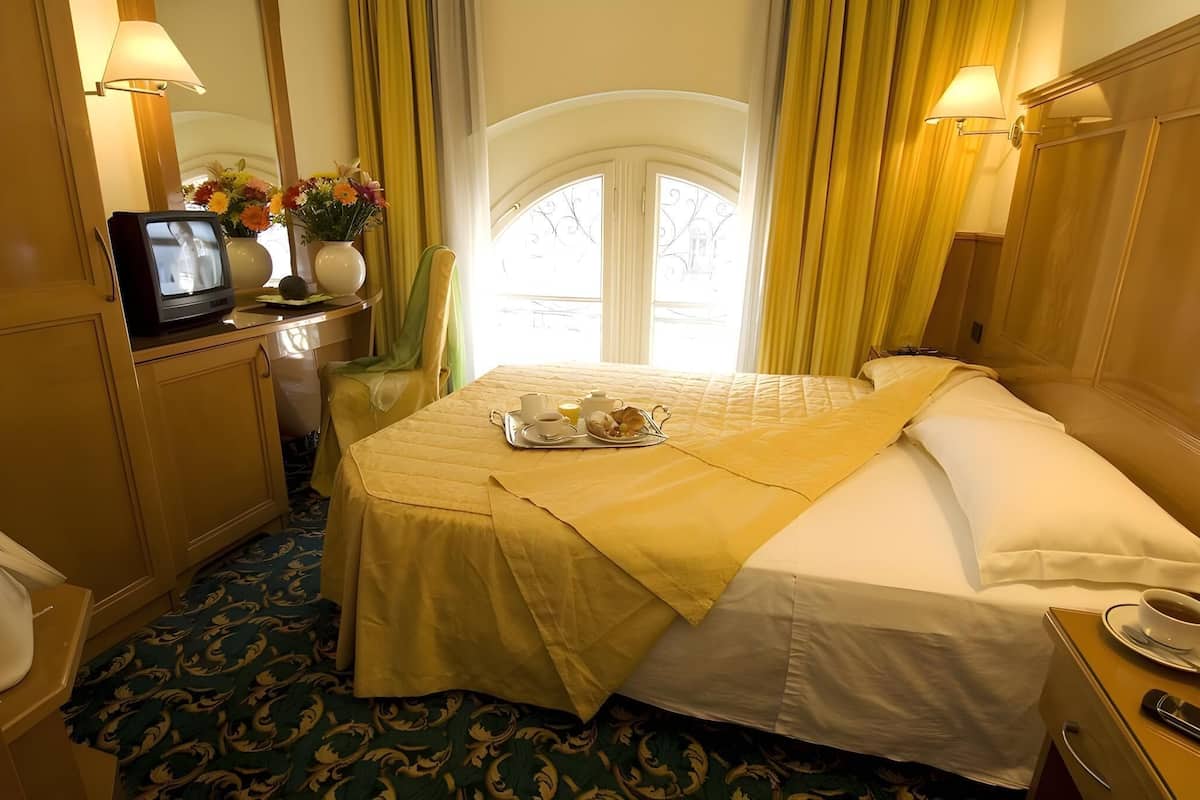 A hotel room with a bed, a desk with a television, a tray with breakfast items, and a window with curtains.