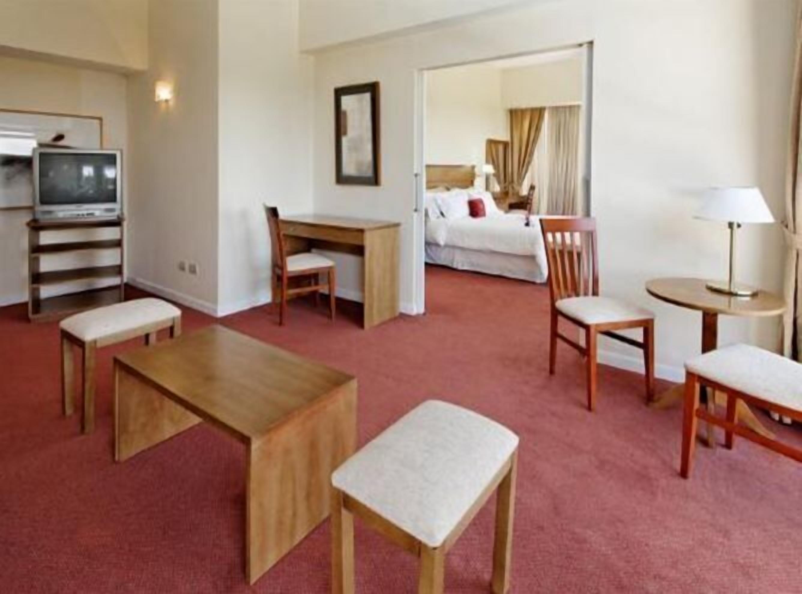A hotel room with a bed, desk, chairs, a television, and a lamp.