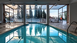Una piscina interior con paredes de vidrio transparente, suelo de ladrillo y vistas a árboles y edificios exteriores.